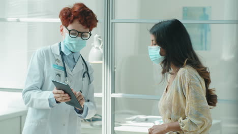 Female-Doctor-and-Patient-in-Masks-Using-Tablet-and-Speaking